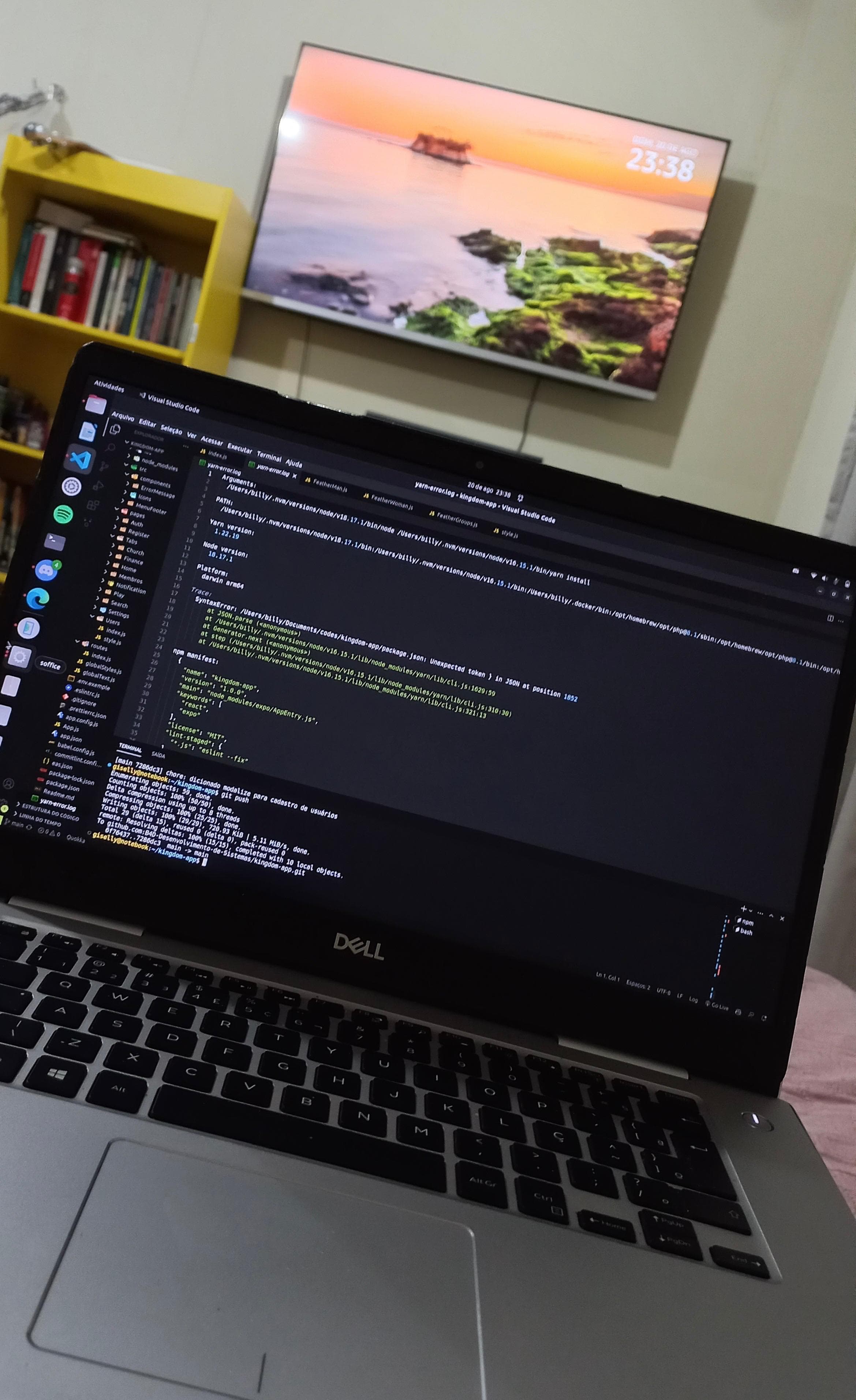 Notebook com o Visual Studio Code aberto, em frente a uma TV e uma estante de livros.